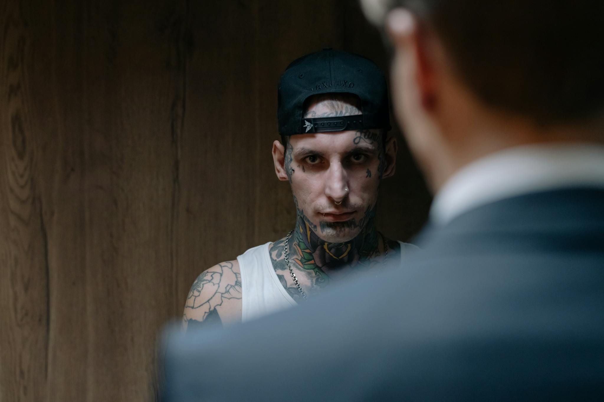 A tattooed man confronts a suited individual during an intense indoor interrogation.