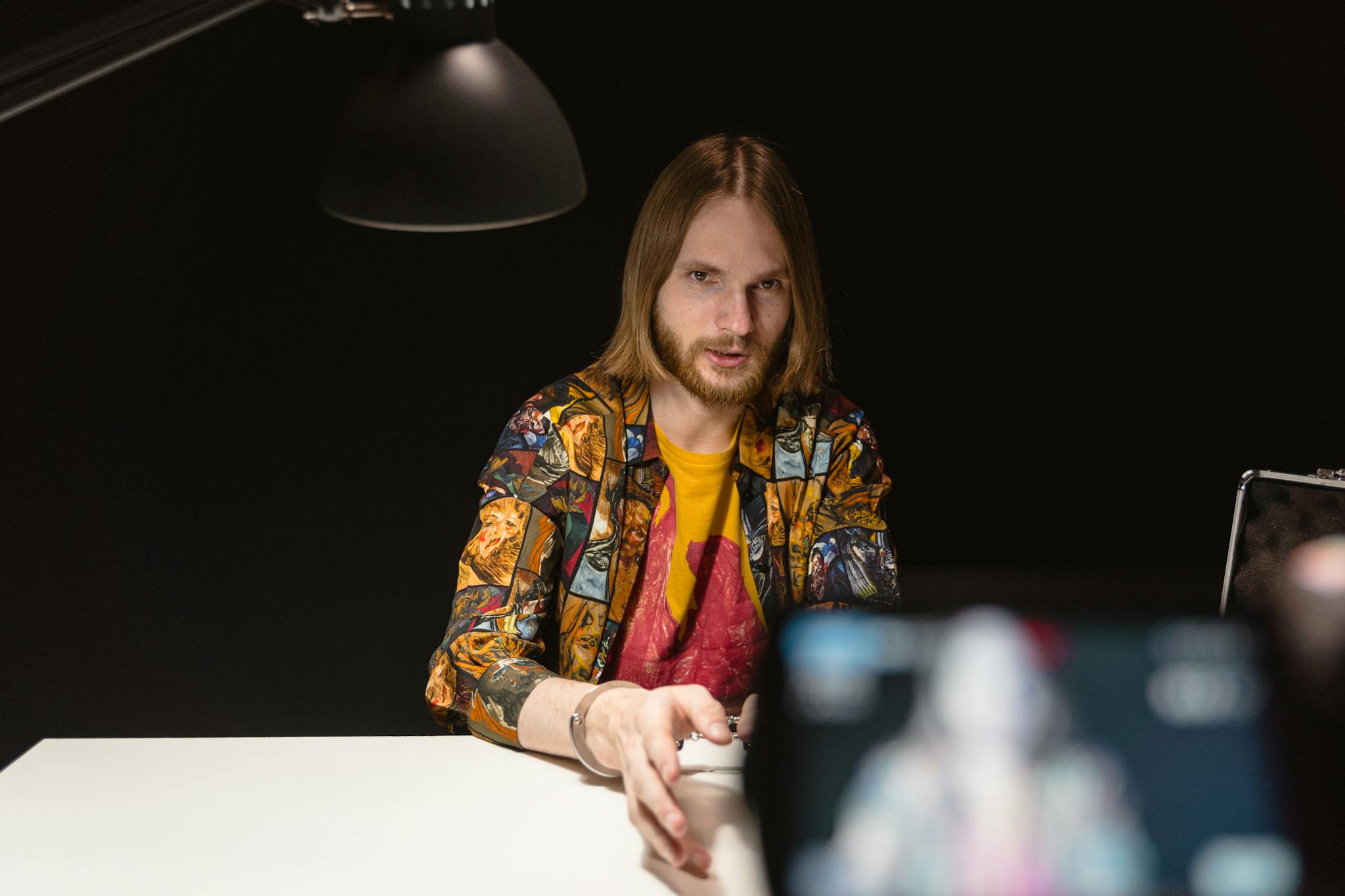 Caucasian man sitting under a lamp in a dark interrogation room setting.